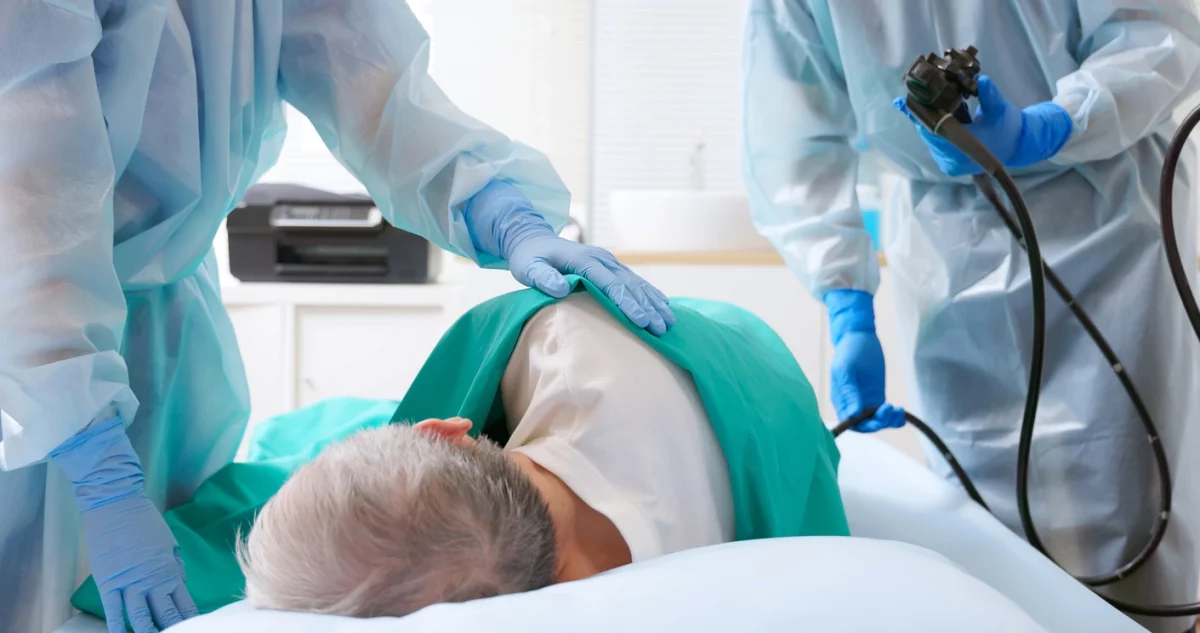 Bild zeigt den Eingriff einer Darmspiegelung in einem medizinischen Rahmen. Patient*in liegt seitwärts auf einer Behandlungsliege. Der Körper ist ab Schulter abwärts mit einem grünen Tuch abgedeckt. An beiden Seiten der Liege steht medizinisches Fachpersonal in Kitteln und Handschuhen. Die rektale Einführung eines Koloskops wird angedeutet. #Darmkrebsvorsroge #Darmspiegelung #Koloskopie
